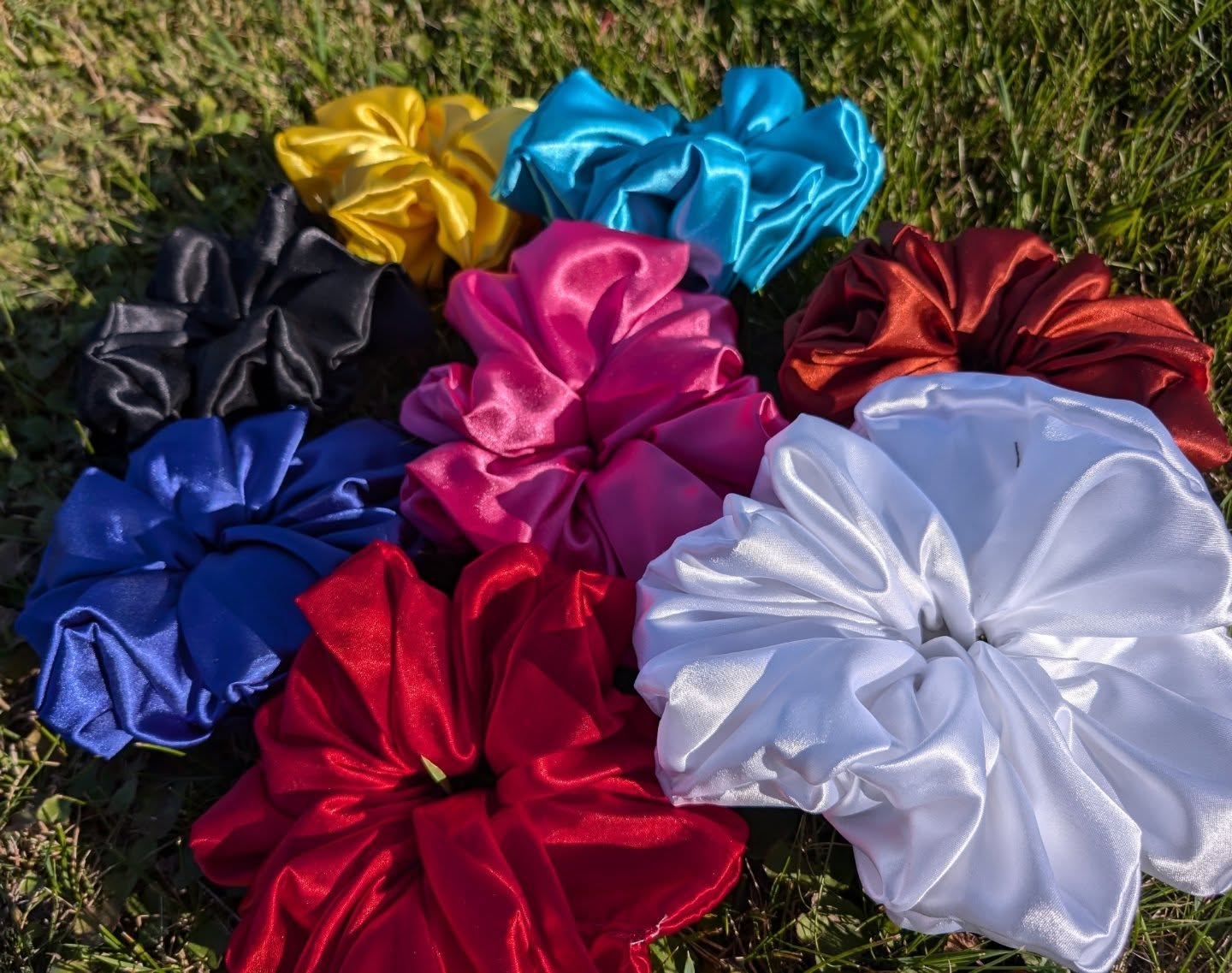 Eight XL (extra large)  satin scrunchies laying on the grass in multiple colors in the sunlight. Yellow, teal, black, pink, copper, royal blue, bright red and white